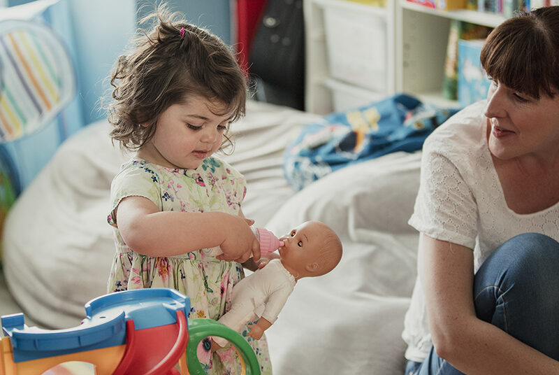 Ruskeatukkainen pieni tyttö leikkii nukella äidin katsoessa vieressä