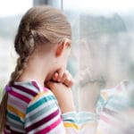 Surullinen kouluikäinen tyttö katselee sateesta märän ikkunan läpi.