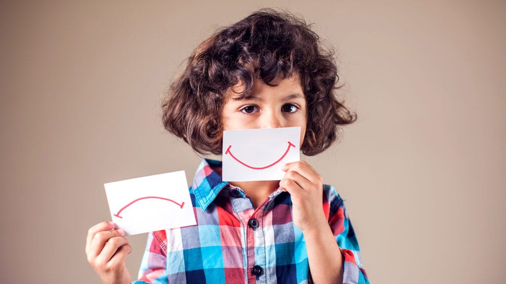 Ruskeatukkainen ja -silmäinen poikalapsi valitsee paperilta hymynaaman tai surunaaman.
