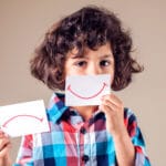 Ruskeatukkainen ja -silmäinen poikalapsi valitsee paperilta hymynaaman tai surunaaman.