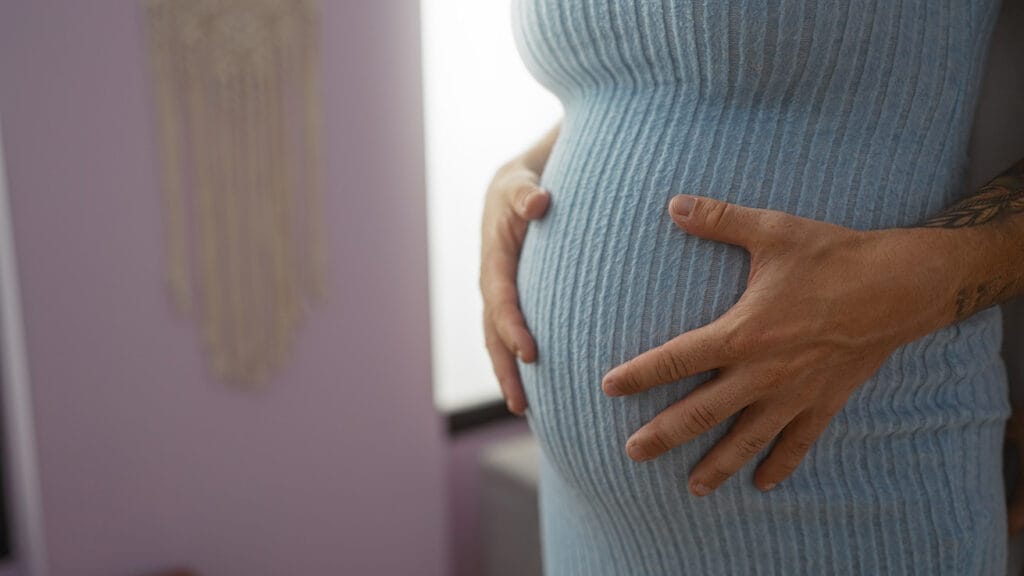 Odottava nainen sinisessä neulemekossa kädet vauvavatsan ympärillä.