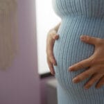 Odottava nainen sinisessä neulemekossa kädet vauvavatsan ympärillä.