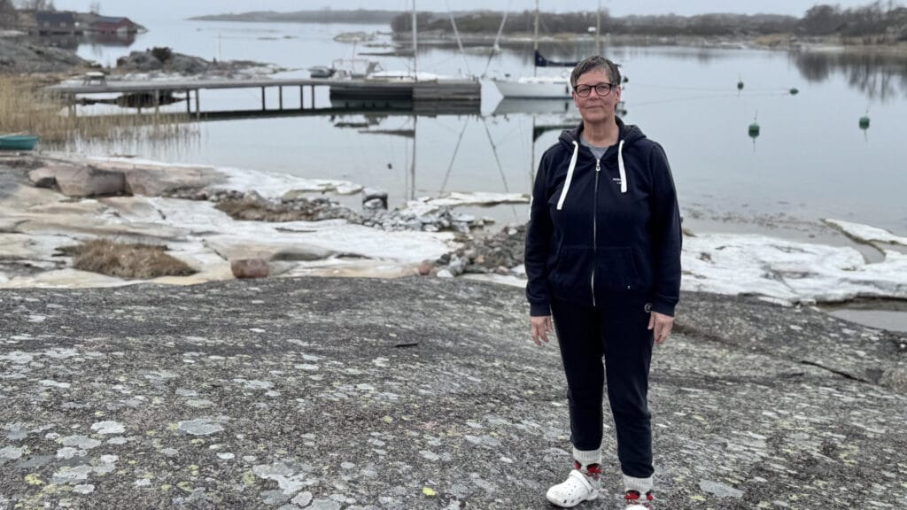 Keski-ikäinen nainen seisoo tumma huppari päällään kalliolla meren rannalla, jonka taustalla näkyy veneitä.