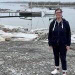 Keski-ikäinen nainen seisoo tumma huppari päällään kalliolla meren rannalla, jonka taustalla näkyy veneitä.