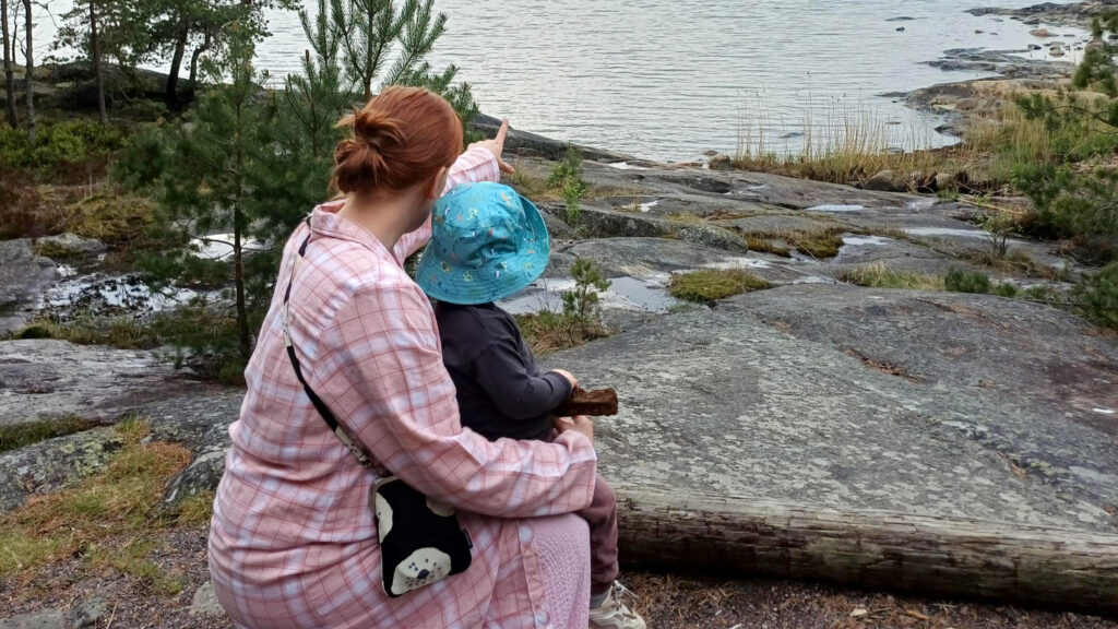 Punatukkainen nainen istuu rantakalliolla pieni sinihattuinen poika sylissään katselee merelle,