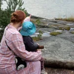 Punatukkainen nainen istuu rantakalliolla pieni sinihattuinen poika sylissään katselee merelle,